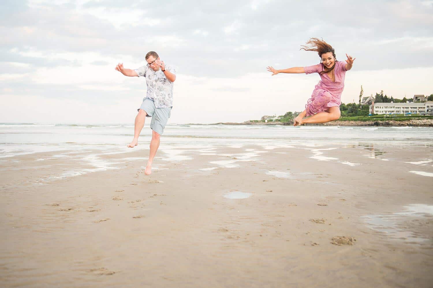 Two people are seen joyfully jumping in the air on a beach, with the sea and buildings in the background.