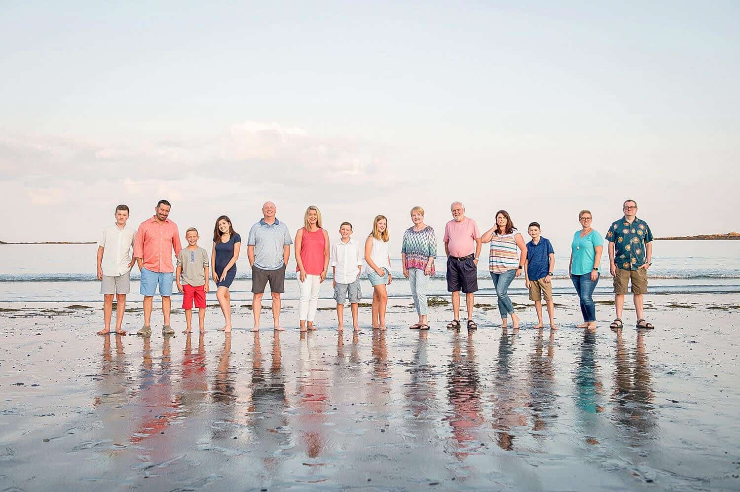 A group of thirteen people of various ages stand in a line on a beach with the ocean in the background. They are dressed in casual summer clothing.