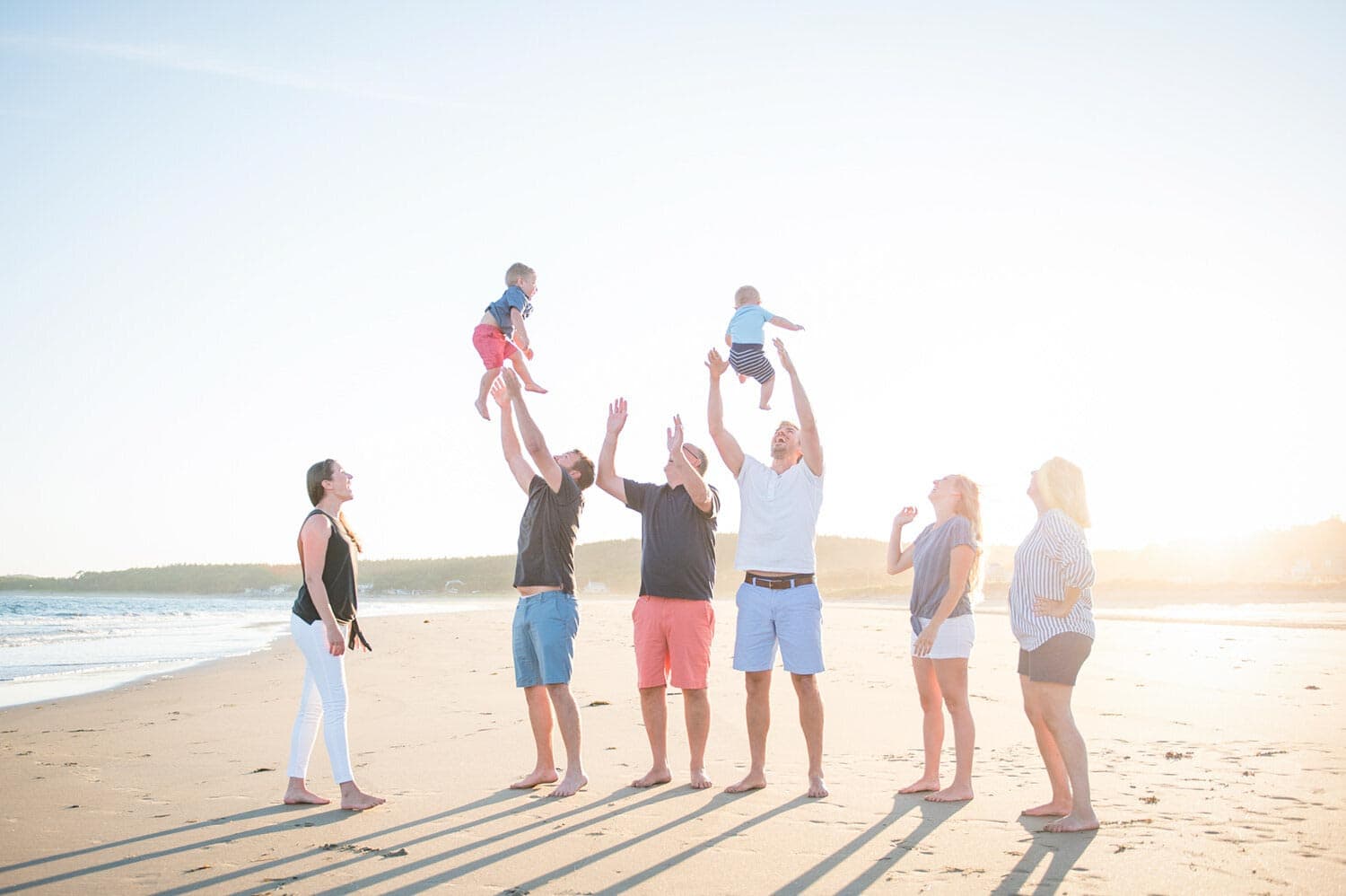 A group of six adults stand on a beach, and two of them are lifting small children into the air. The sky is clear and the sun is bright.