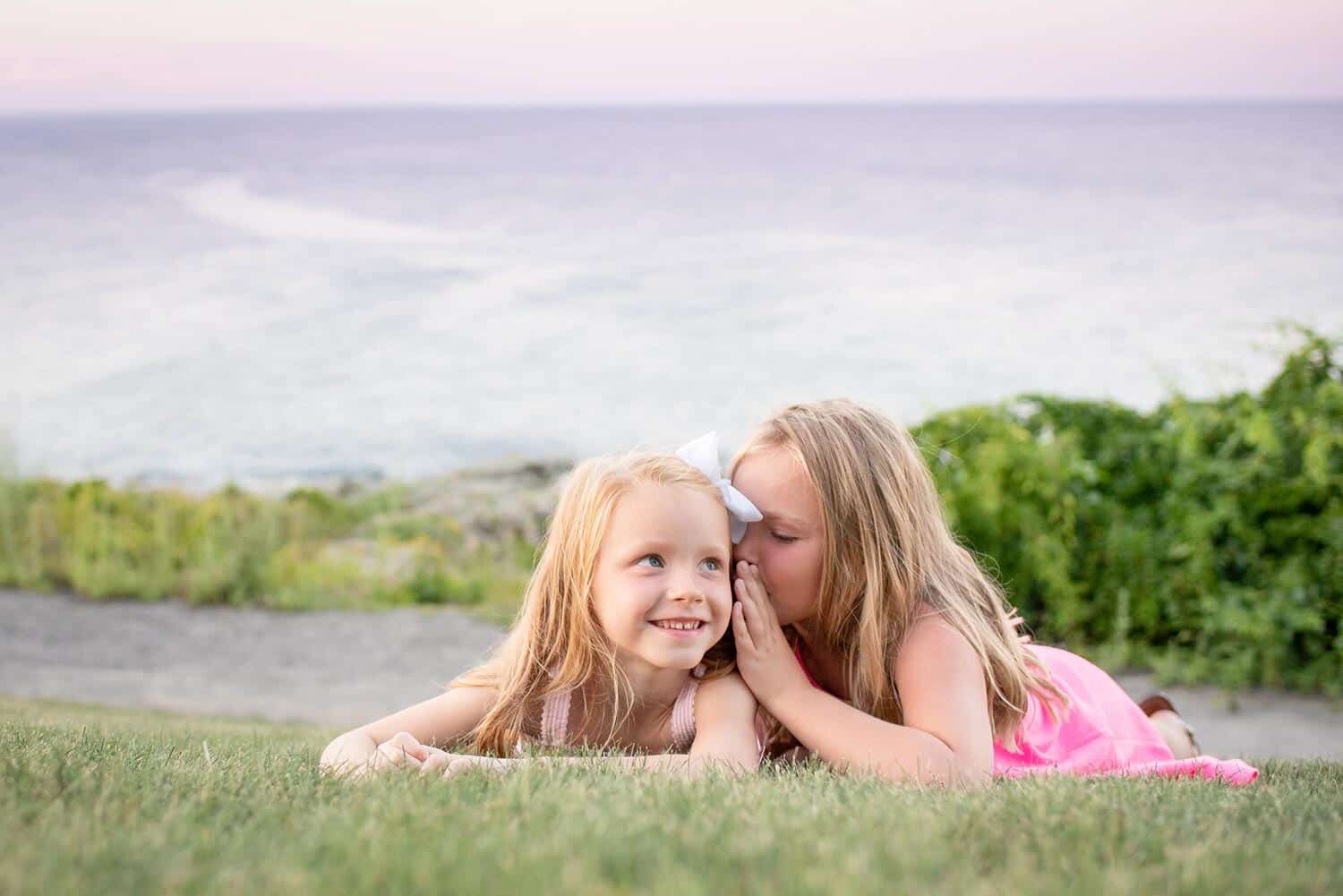 Two young girls lying on grass with an ocean in the background. One girl is whispering into the other girl's ear, who is smiling and looking ahead.