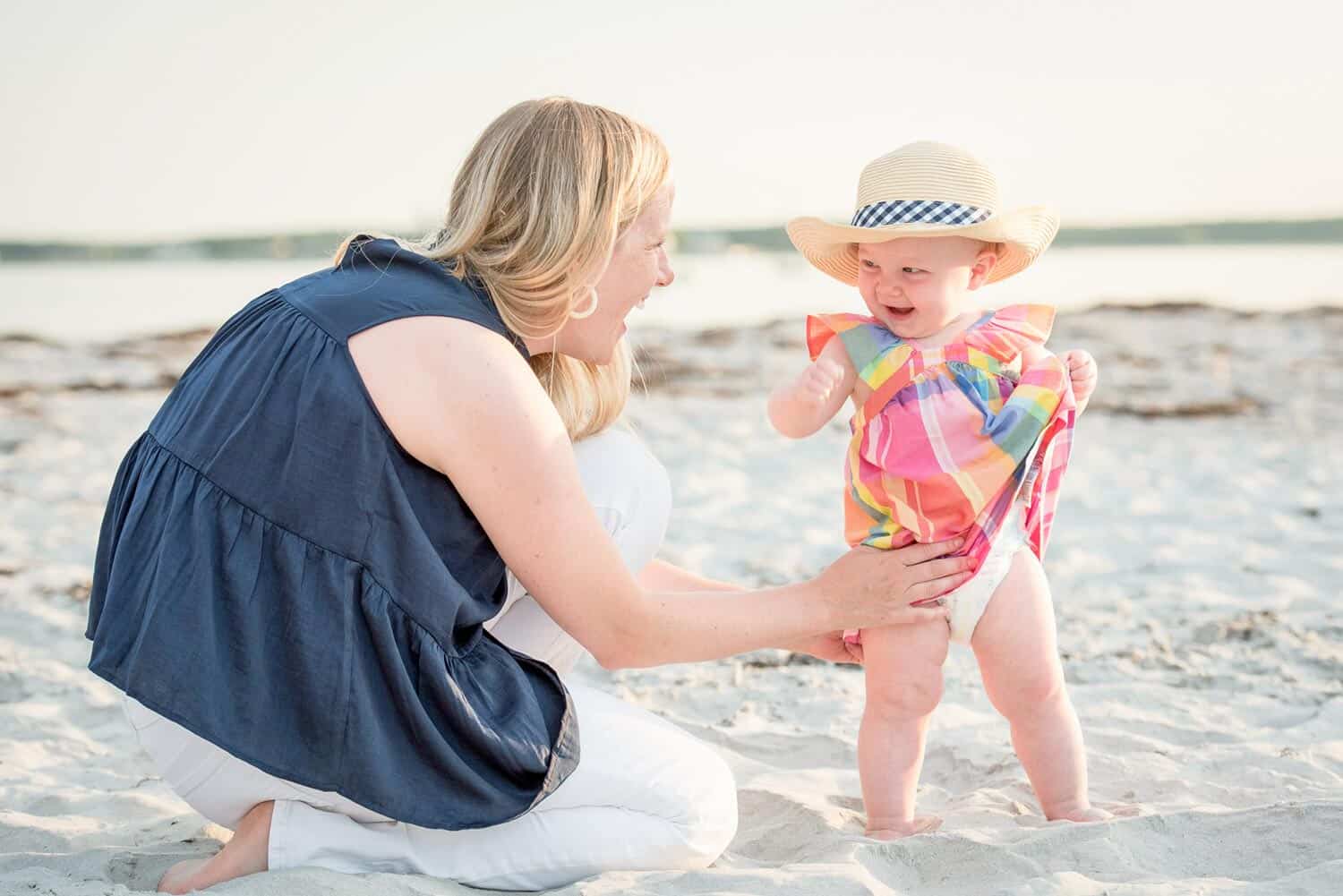 A woman and a baby in a colorful outfit and straw hat are playing on a sandy beach. The woman is kneeling and smiling at the standing baby.