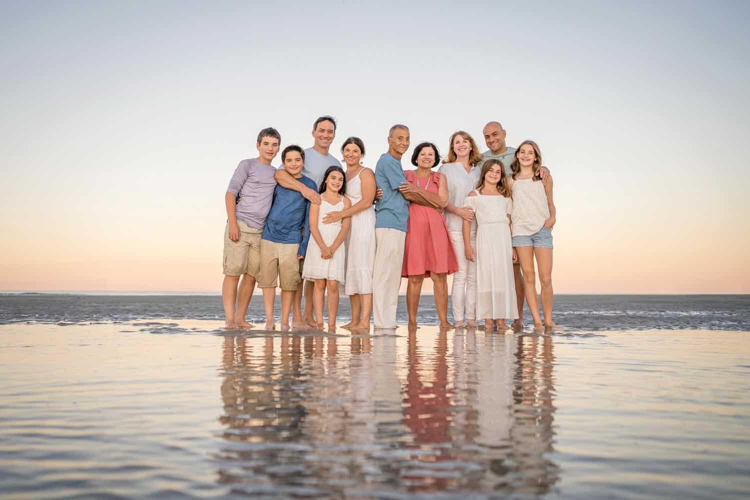 A group of people on the beach, their reflections visible on the wet sand. They are smiling and dressed in casual summer clothing. The sky is clear.