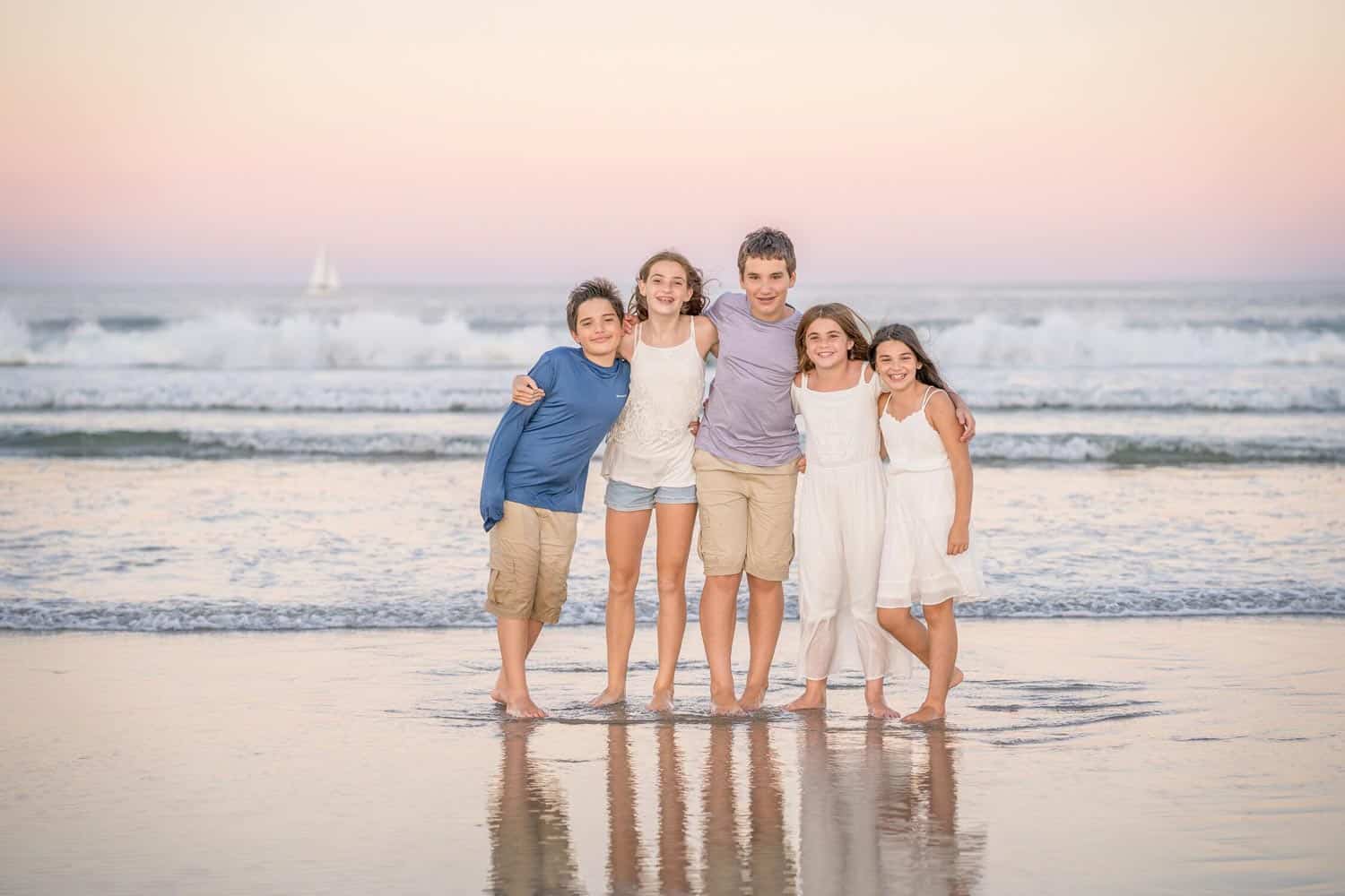 5 children stand on a beach with their feet in the water at sunset. Waves crash in the background, and a sailboat is visible on the horizon.