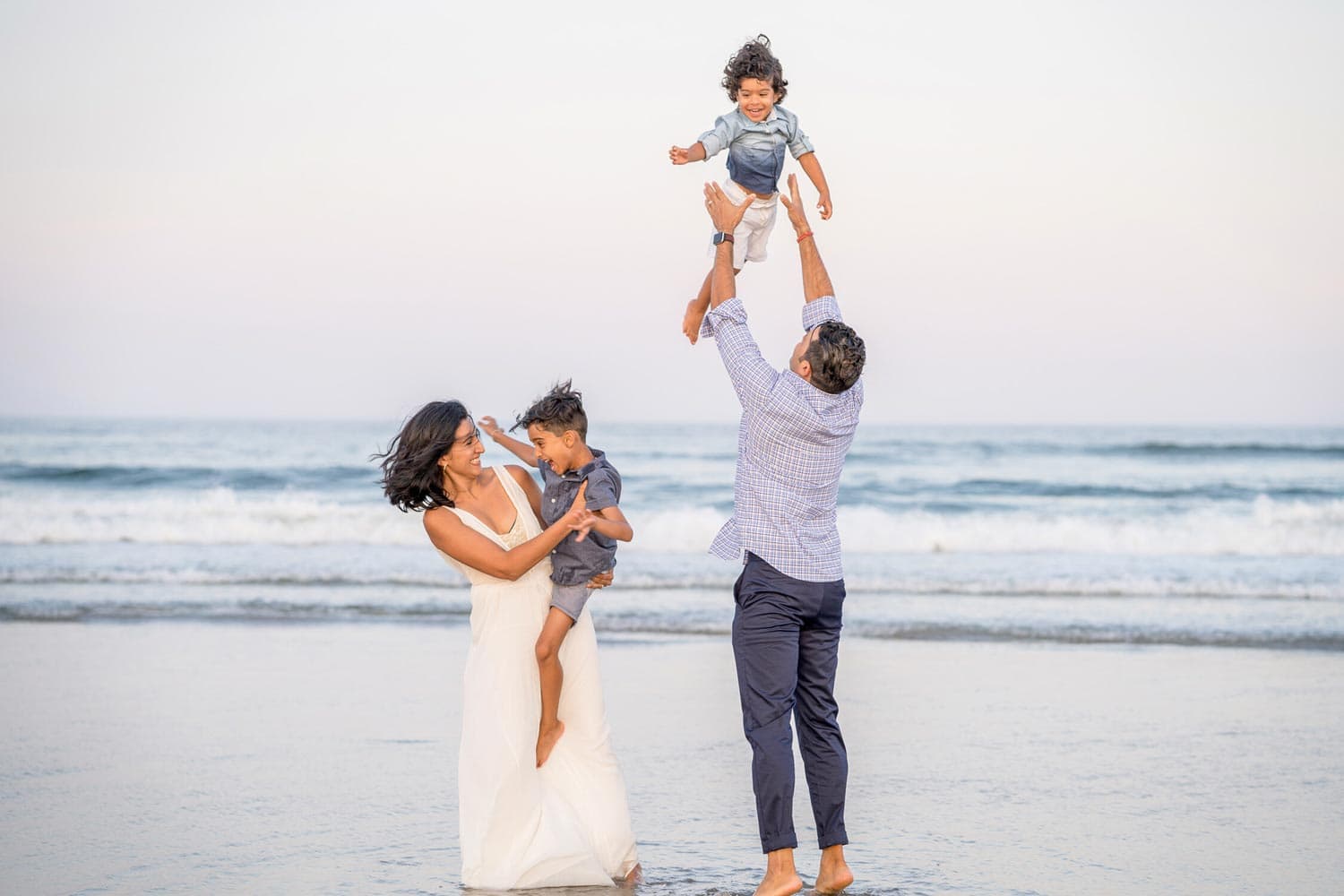 A family of four plays at the beach. The mother holds one child while the father lifts the other child above his head.