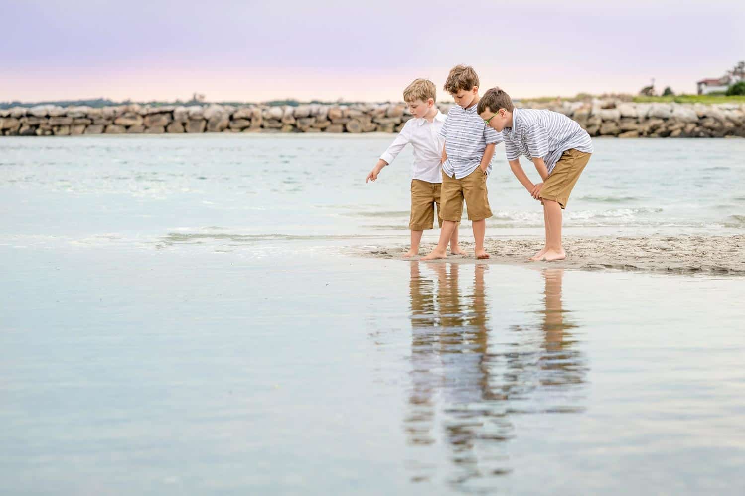 Three boys in similar outfits stand barefoot on a sandy beach, peering into the shallow water and pointing at something near the shore.