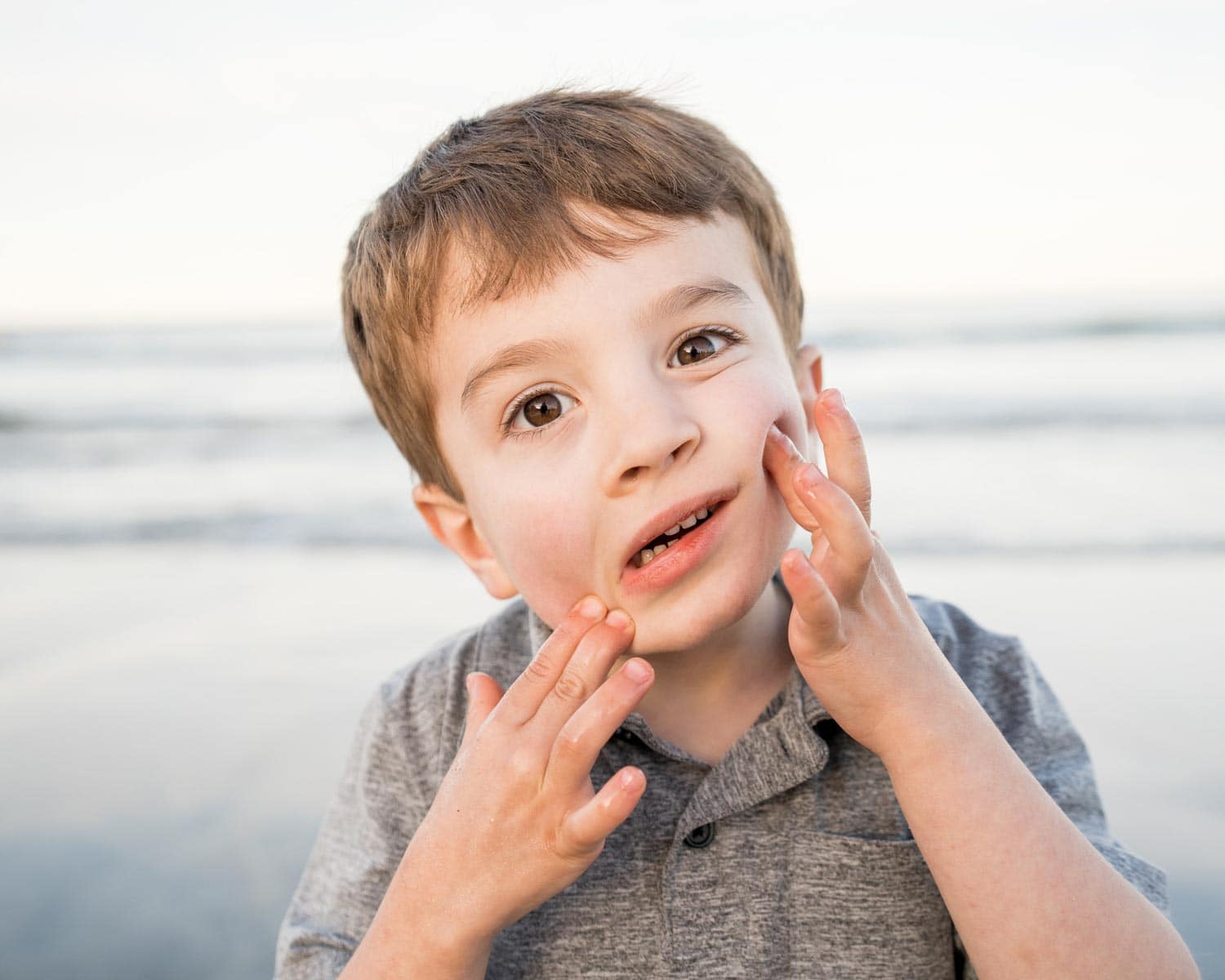 Young child with short brown hair touches his cheeks while standing on a beach with the ocean in the background.