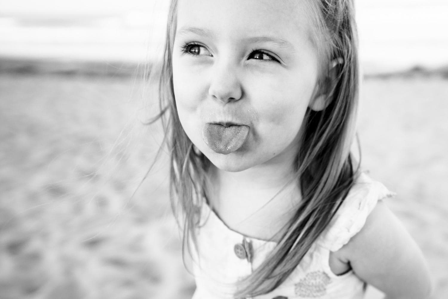 Black and white photo of a young girl with long hair sticking out her tongue while standing on a beach. She wears a tank top and looks slightly to the side.