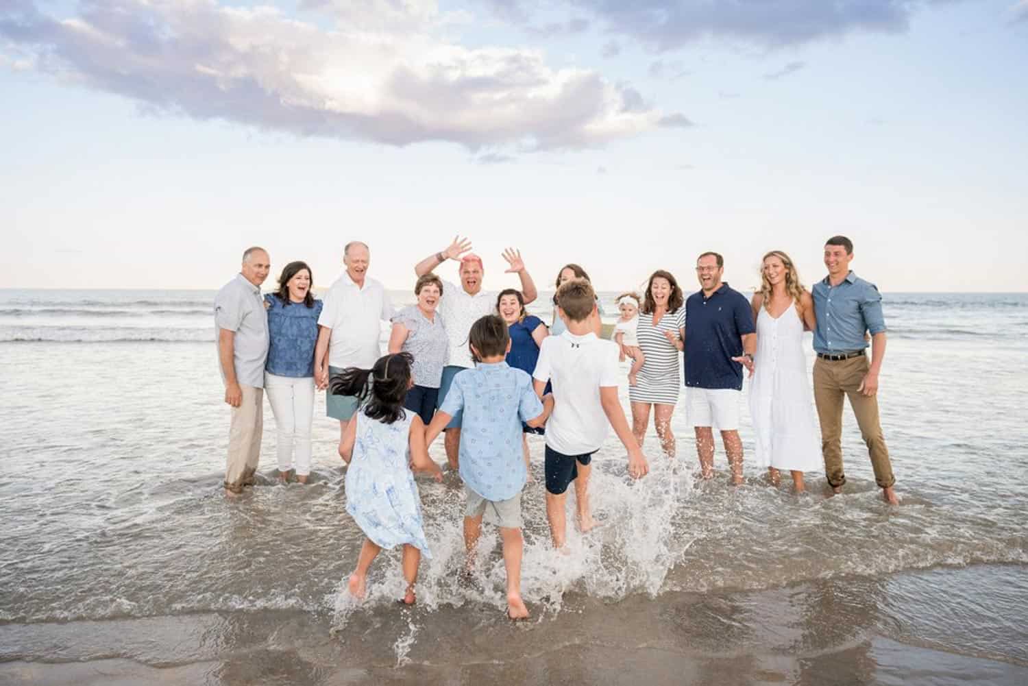 A group of people stands together in shallow ocean water, laughing and waving as four children splash in front of them. The sky is partly cloudy.