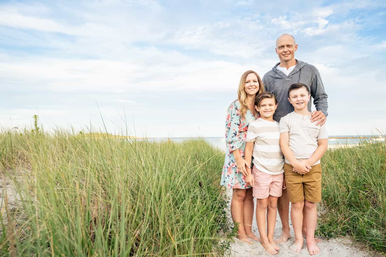 A family of 4 on a beach with grassy dunes, smiling at the camera. The mother is wearing a floral dress, and the father and two boys are wearing casual summer clothes.