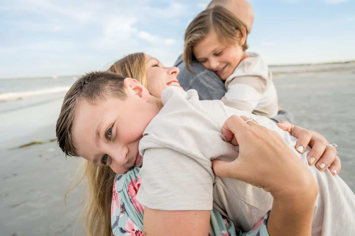 A family of 4 shares a group hug on the beach. The mother holds a young boy smiling at the camera, while the father holds another child in the background.