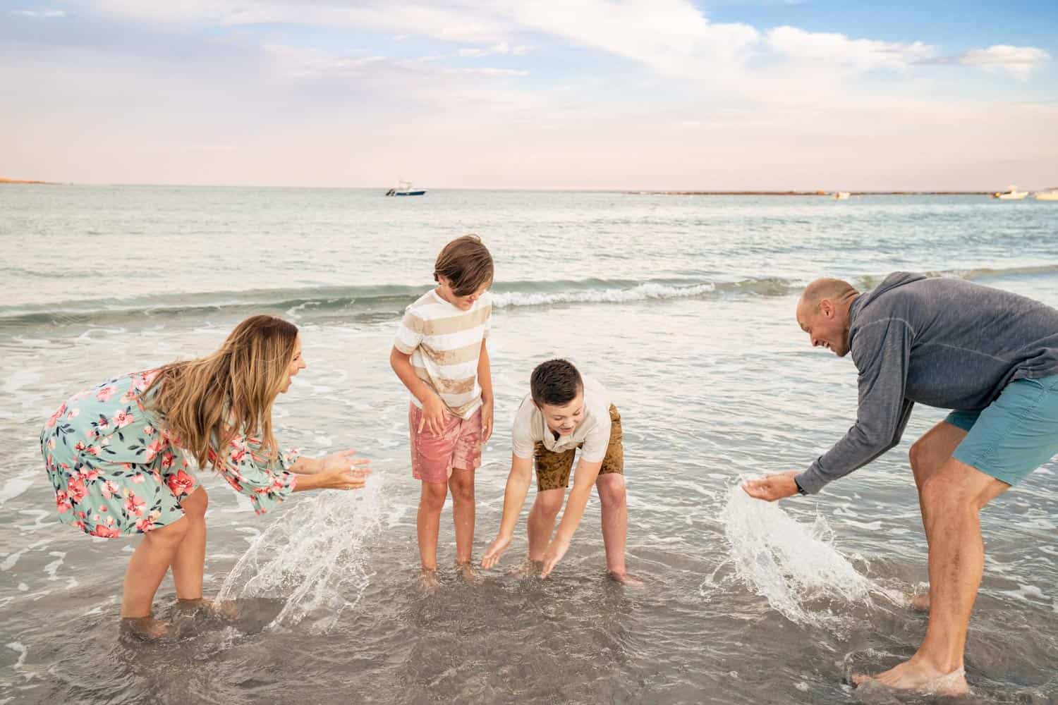 A family of four playing in the shallow water at the beach; two adults and two children are splashing water at each other.
