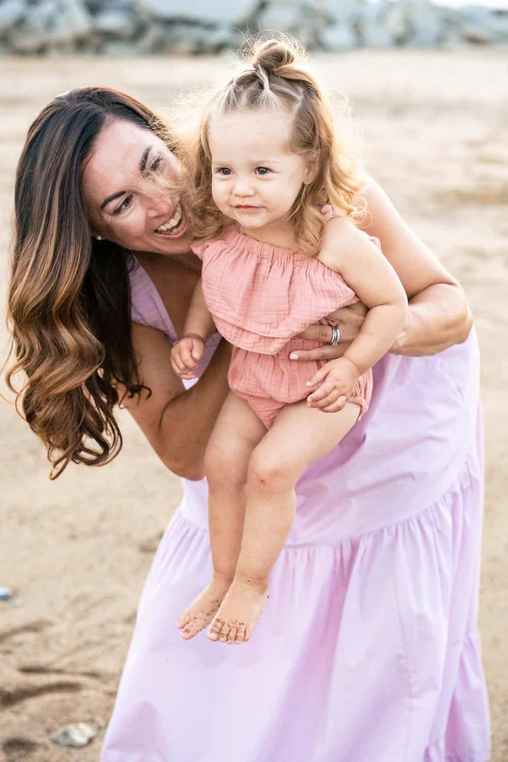 A woman in a pink dress holds a young child in a pink outfit while they both smile on a sandy beach.