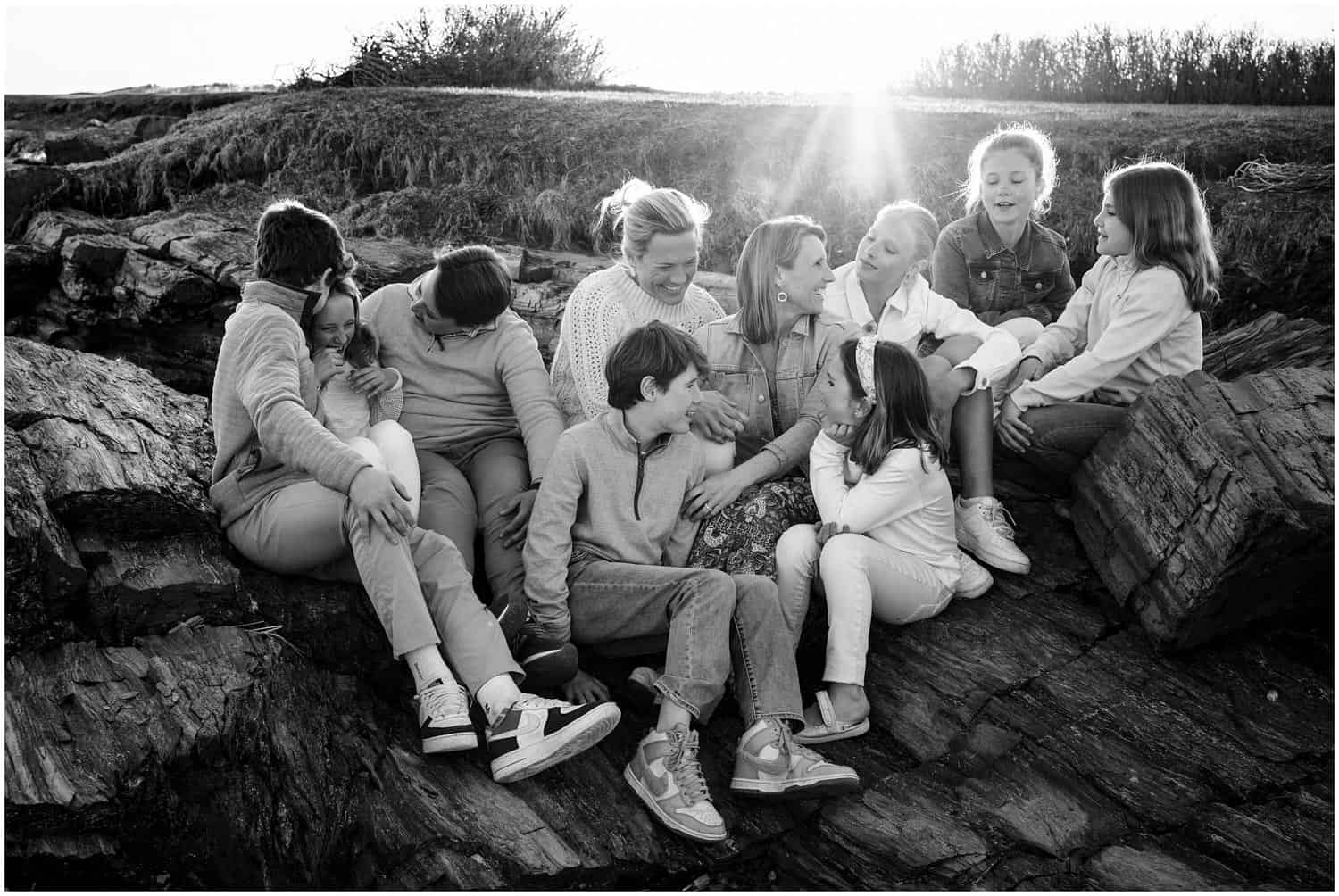 A group of children and adults, seated on a rocky surface outdoors, are smiling and interacting with each other. The sun is shining brightly in the background.