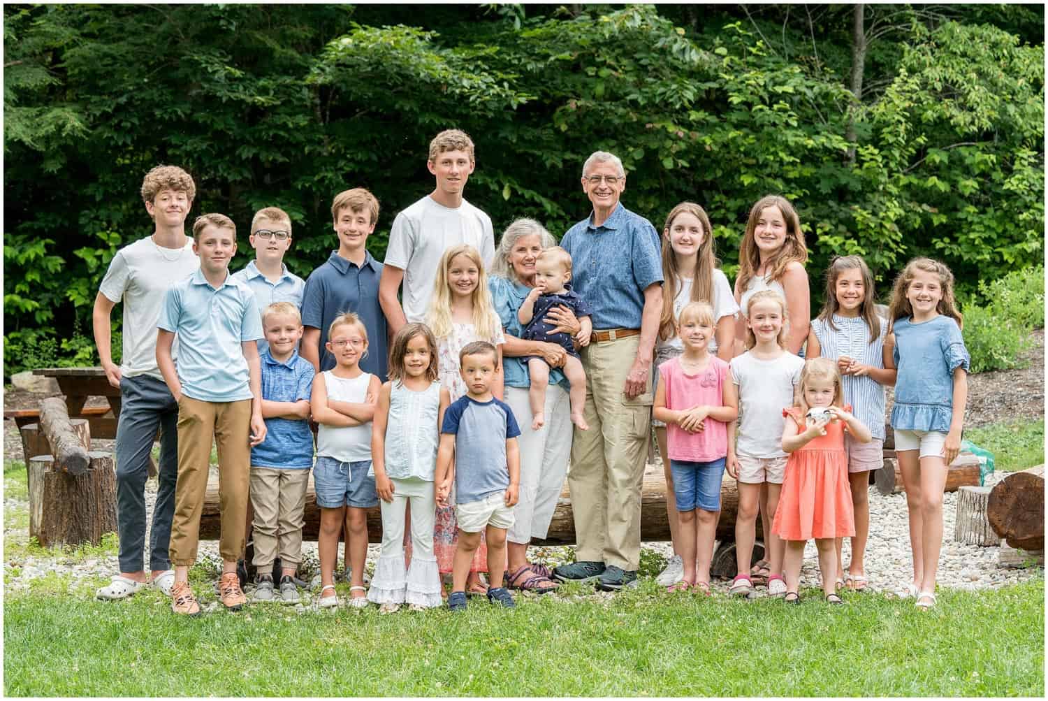 A group of various adults and children, stands together outdoors on grass with trees in the background. They are all looking at the camera and smiling.