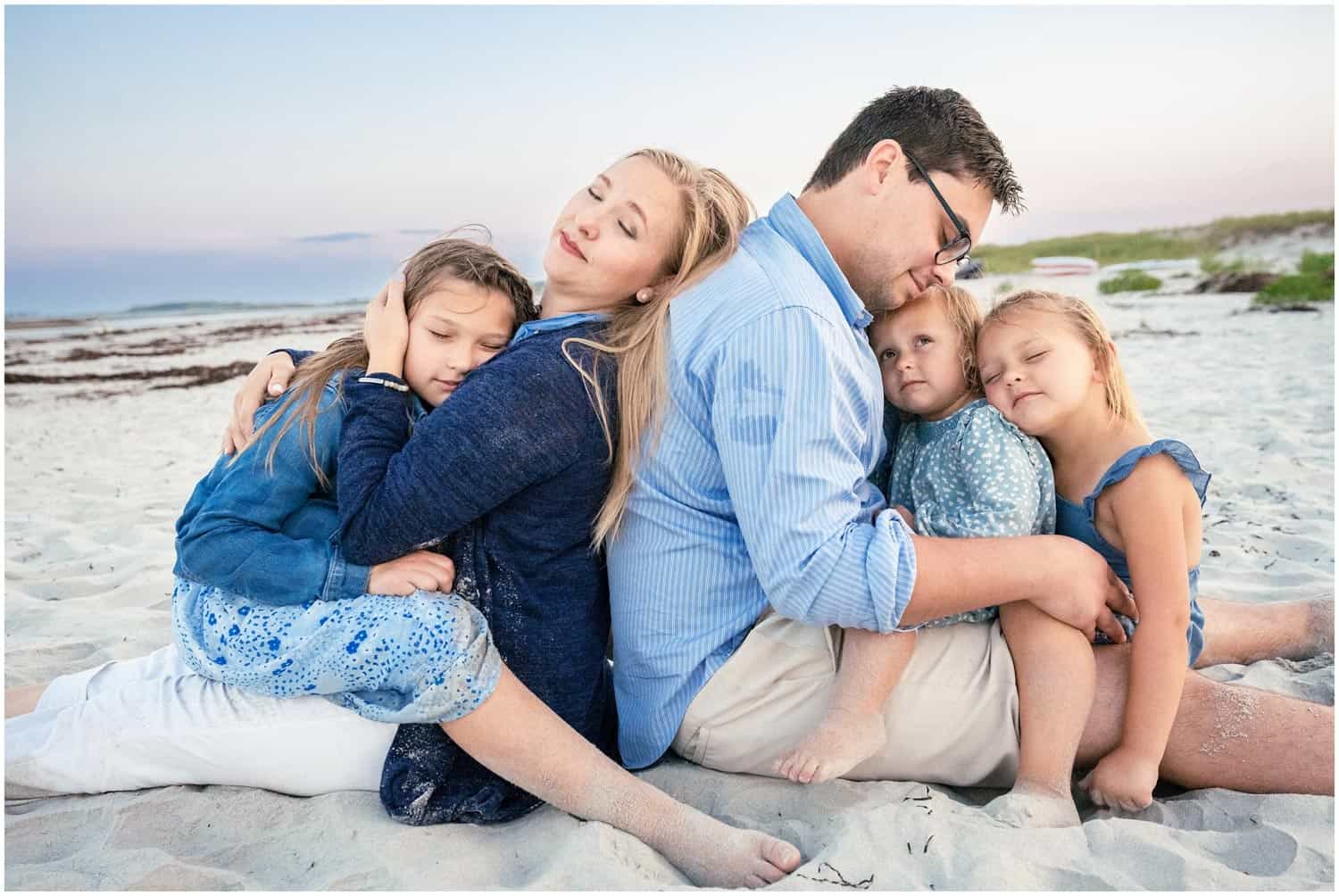 A man and woman sit back-to-back on a beach, hugging their children. The group appears relaxed and content. The beach and ocean are visible in the background.