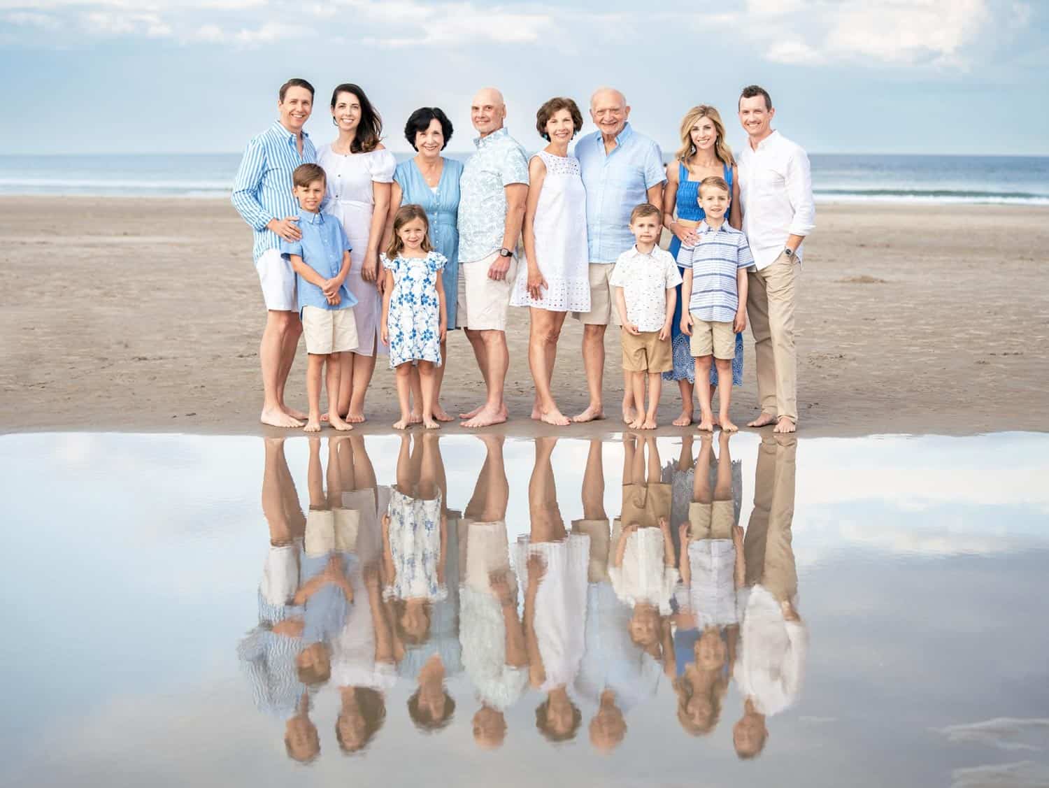 A group of adults and children stand on a beach in front of a shallow water pool, reflecting their image. The beach and sea are visible in the background.