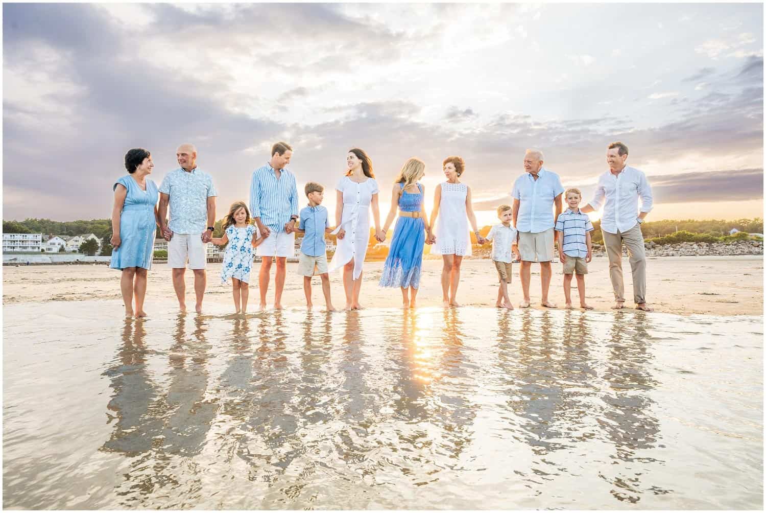 A group of 11 children and adults, stand in shallow water on a beach holding hands. They are dressed in casual summer clothing with houses in the background.