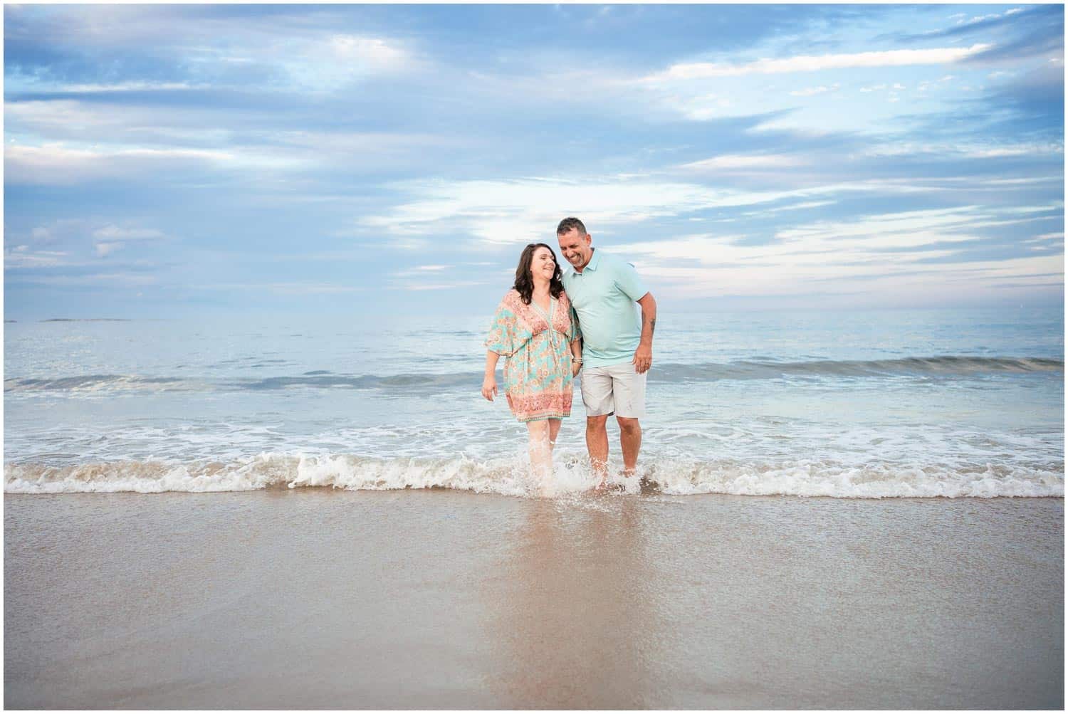 A couple walks along the shoreline, feet in the water, under a partly cloudy sky.