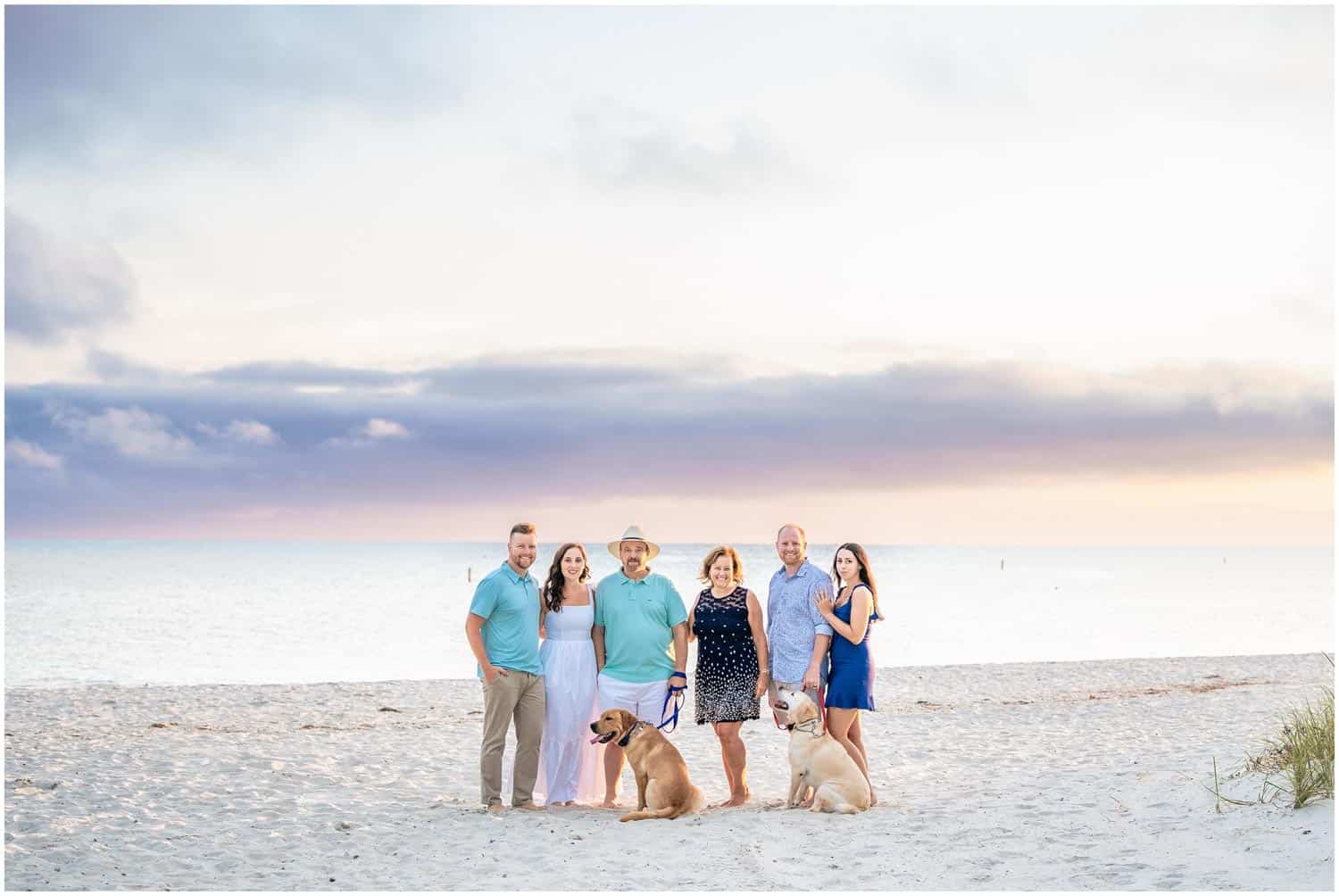 A group of six adults and two dogs stand on a beach at sunset, with the ocean and a cloudy sky in the background.