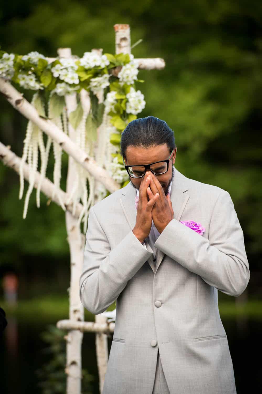 A man in a light gray suit and glasses stands with his hands clasped together, appearing contemplative in front of a floral wedding arch outdoors.