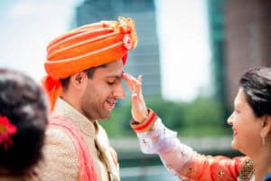 A man wearing a traditional turban is being blessed by a woman touching his forehead. The background shows tall buildings and greenery.