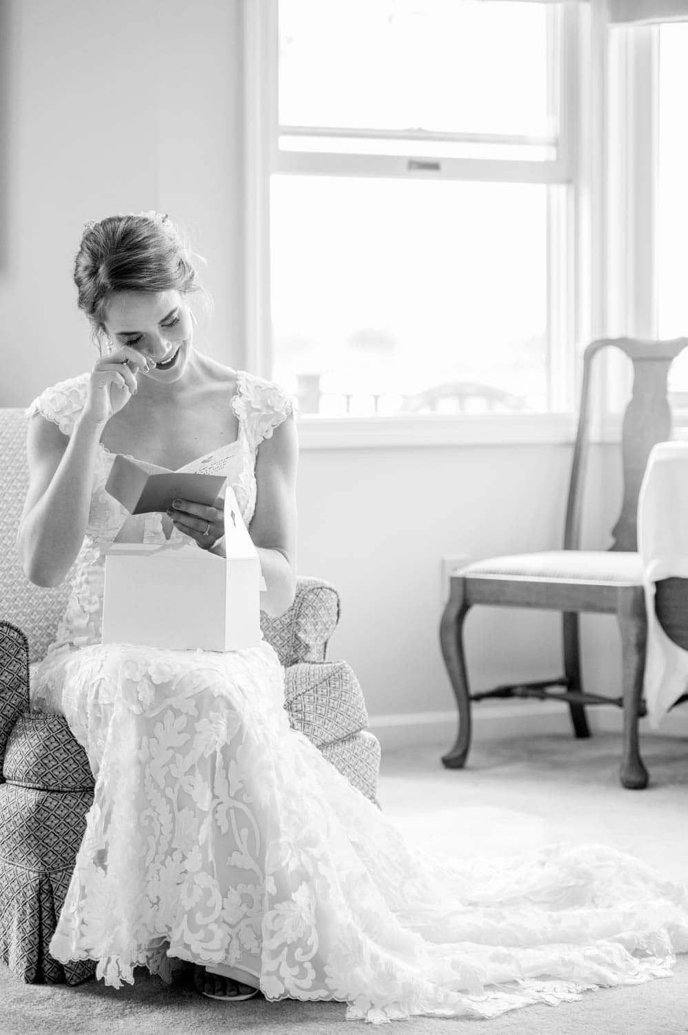 A bride in a lace gown sits in a chair by a window, smiling and touching her face while holding a card and a box.