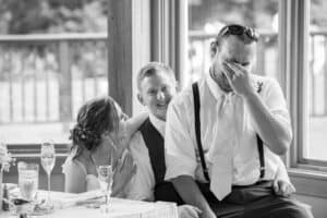 2 people in wedding attire sit at a table, while a groom dressed in a shirt and suspenders sits on the man's lap, covering his face with one hand and laughing.