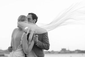A bride and groom share a kiss while a veil flows in the wind in a black and white photo. The bride's back is to the camera, and they are outdoors.
