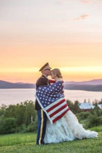 A couple stands embracing at sunset, both wrapped in an American flag. The groom is in a military uniform, and the bride is in a white wedding dress.