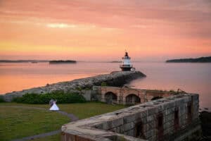 A bride and groom by the water with a stone path leading to a lighthouse at sunset. The sky is filled with warm colors, and the sea stretches to the horizon.