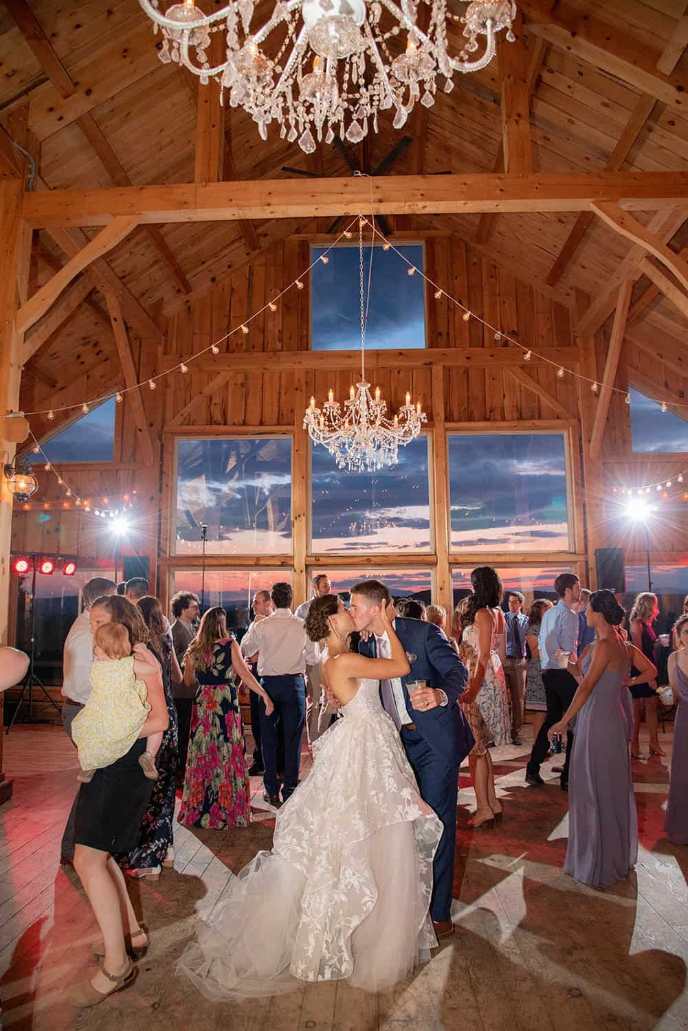 A couple in wedding attire shares a dance in a wooden hall with chandeliers, surrounded by guests, some dancing and others observing.