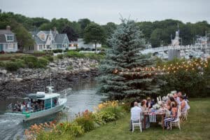 Group of people seated at a long table set for a meal by a riverbank with a nearby docked boat. Houses and trees are in the background, and string lights hang above.