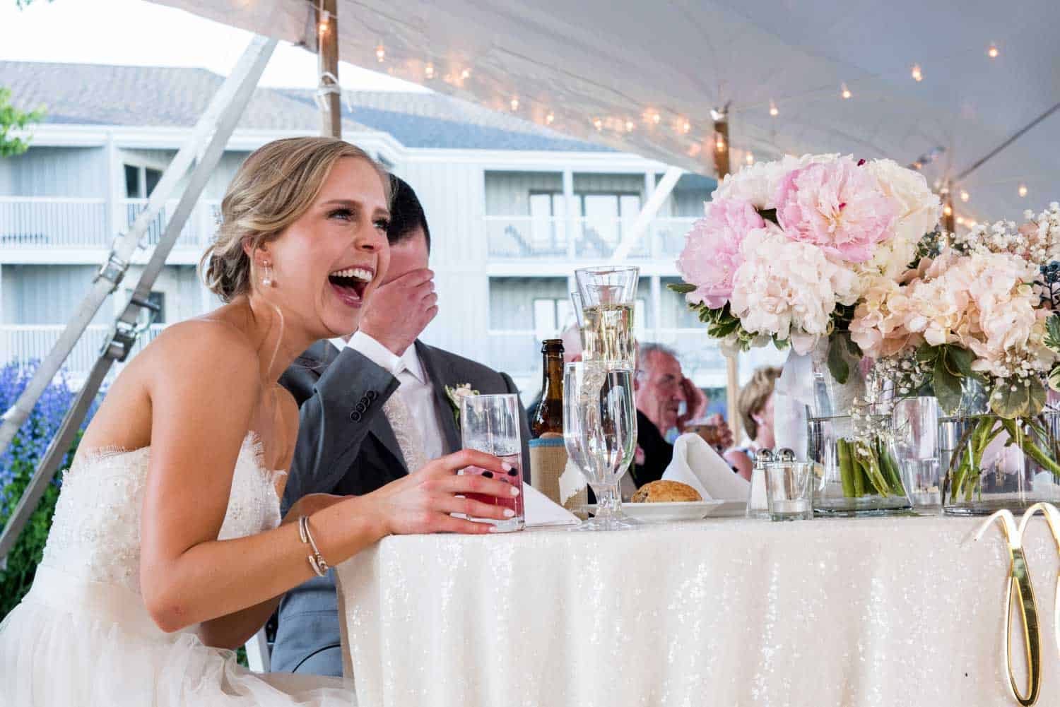 A bride in a white dress laughs while seated at a table with a groom in a suit covering his face. The table has drinks, flowers, and plates.