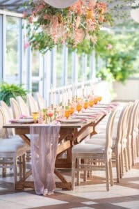 A long dining table is set with pink and orange glassware, white candles, and a sheer pink table runner. A floral arrangement with pink and green foliage hangs overhead.