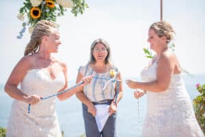 Two women in wedding dresses hold either end of a rope in front of an officiant by the seaside. A floral arch of sunflowers and white flowers frames the scene.