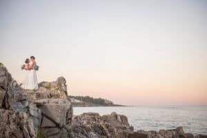 Two brides in white gowns stand on rocky cliffs by the sea at sunset, holding bouquets and embracing against a pastel sky backdrop.