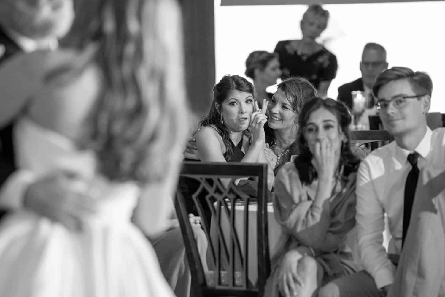 A black-and-white photo shows guests at a wedding reception reacting emotionally, some wiping tears and others smiling, as they watch the couple's first dance.