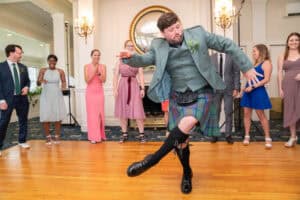 A man in a kilt energetically dances on a wooden floor while several people in formal attire watch and smile in the background.