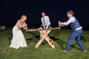 A bride, a groom, and a man in formal attire are using a large saw to cut a log outdoors at night. The log is supported by a wooden stand.