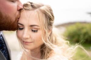Close-up of a man kissing a woman's forehead outdoors. The woman has closed eyes and light hair, while the man has a beard. A blurred natural background is visible.