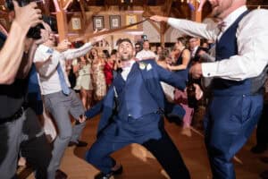 A man in a blue suit dances energetically in the center of a joyous crowd at a lively wedding reception.