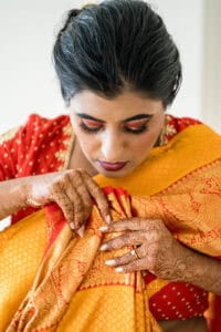 A woman with henna-decorated hands adjusts a yellow and red saree. She has dark hair and is wearing makeup including bold eyeshadow and lipstick.
