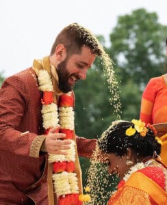A man is pouring rice over a woman's head during a traditional wedding ceremony. Both are wearing ornate, colorful attire and garlands.