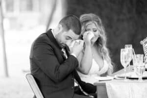 A bride and groom sit at a table, both wiping tears from their eyes, during a wedding reception.