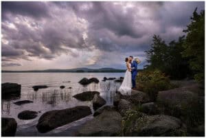 A couple in wedding attire stands on large rocks by a lake under a dramatic, cloudy sky with distant mountains in the background.