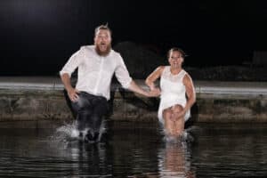 A man and a woman, dressed in formal attire, are holding hands and jumping into the water. They are both smiling and appear to be enjoying the moment.