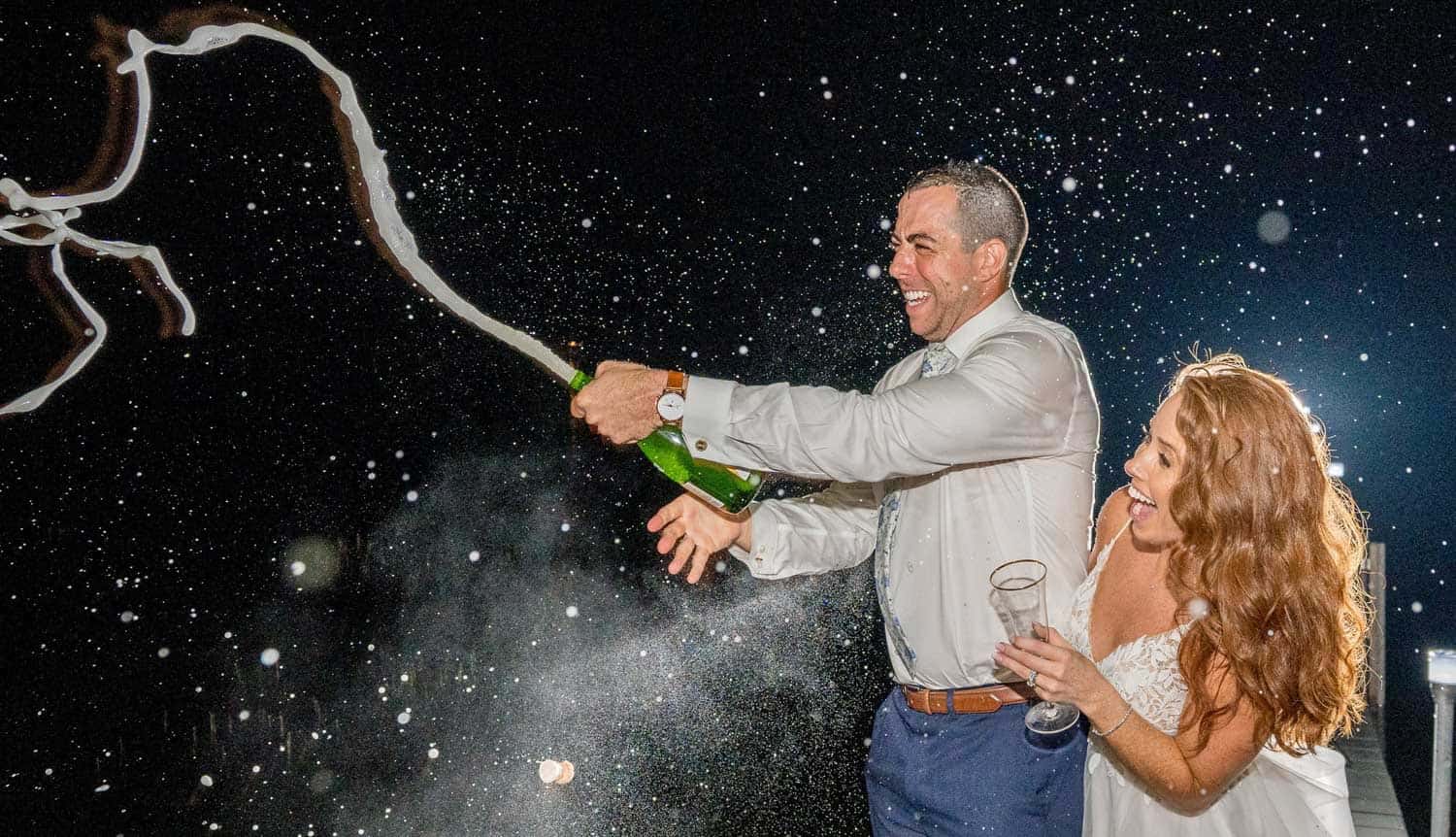 A man in a white shirt pops a champagne bottle, causing it to spray, while a woman in a white dress holding a glass smiles beside him.