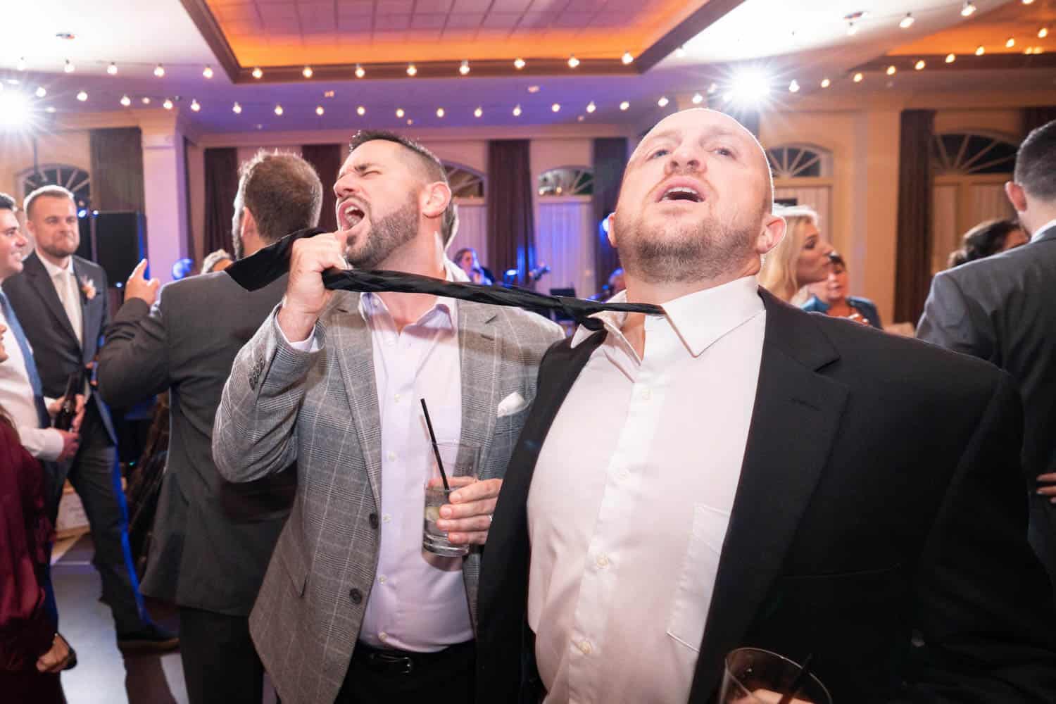 Two men in suits are singing passionately at a reception. One man is tugging on the other's tie while holding a drink, surrounded by other people in party attire.