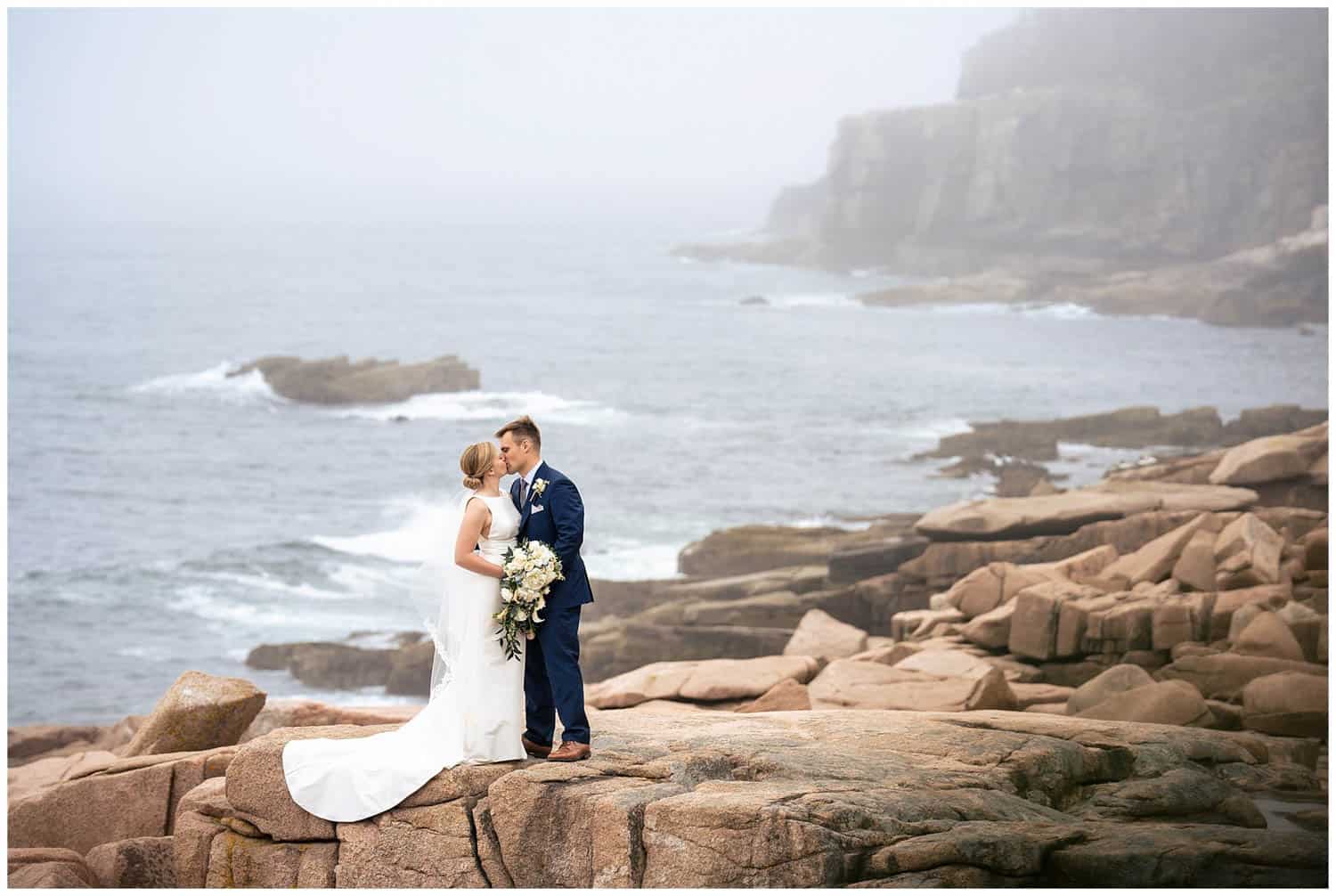 A couple dressed in wedding attire stands on a rocky coastline, sharing a kiss with the ocean and misty cliffs in the background. The bride holds a bouquet of flowers.