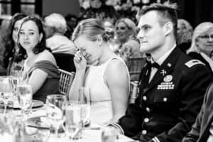A bride wipes her eyes while seated at a dining table next to a groom in military attire and another woman in an off-shoulder dress.